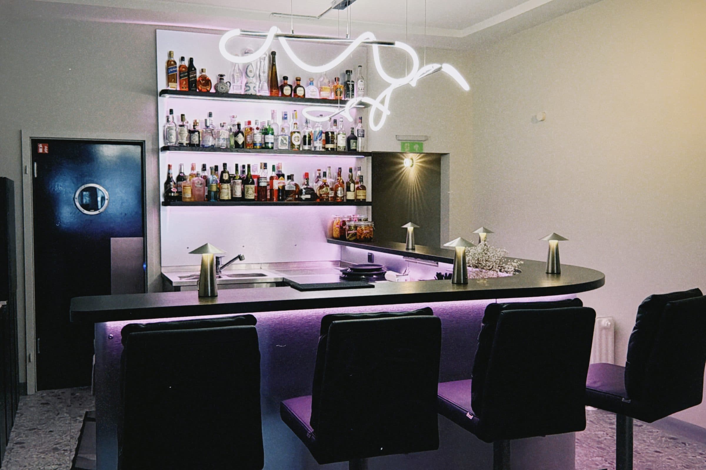 Die Bar des ROMI Studio: geschwungene Edelstahltheke mit schwarzen Barhockern, beleuchtetes Flaschenregal und Neon-Schriftzug an der Wand