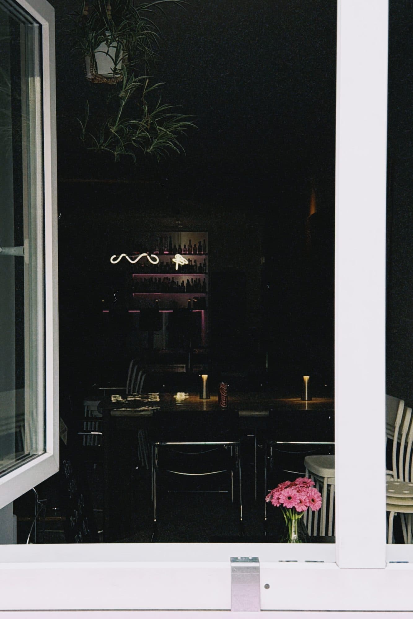 Blick durchs offene Fenster ins abends beleuchtete ROMI Studio, Neon-Schriftzug und Blumen auf dem Tisch
