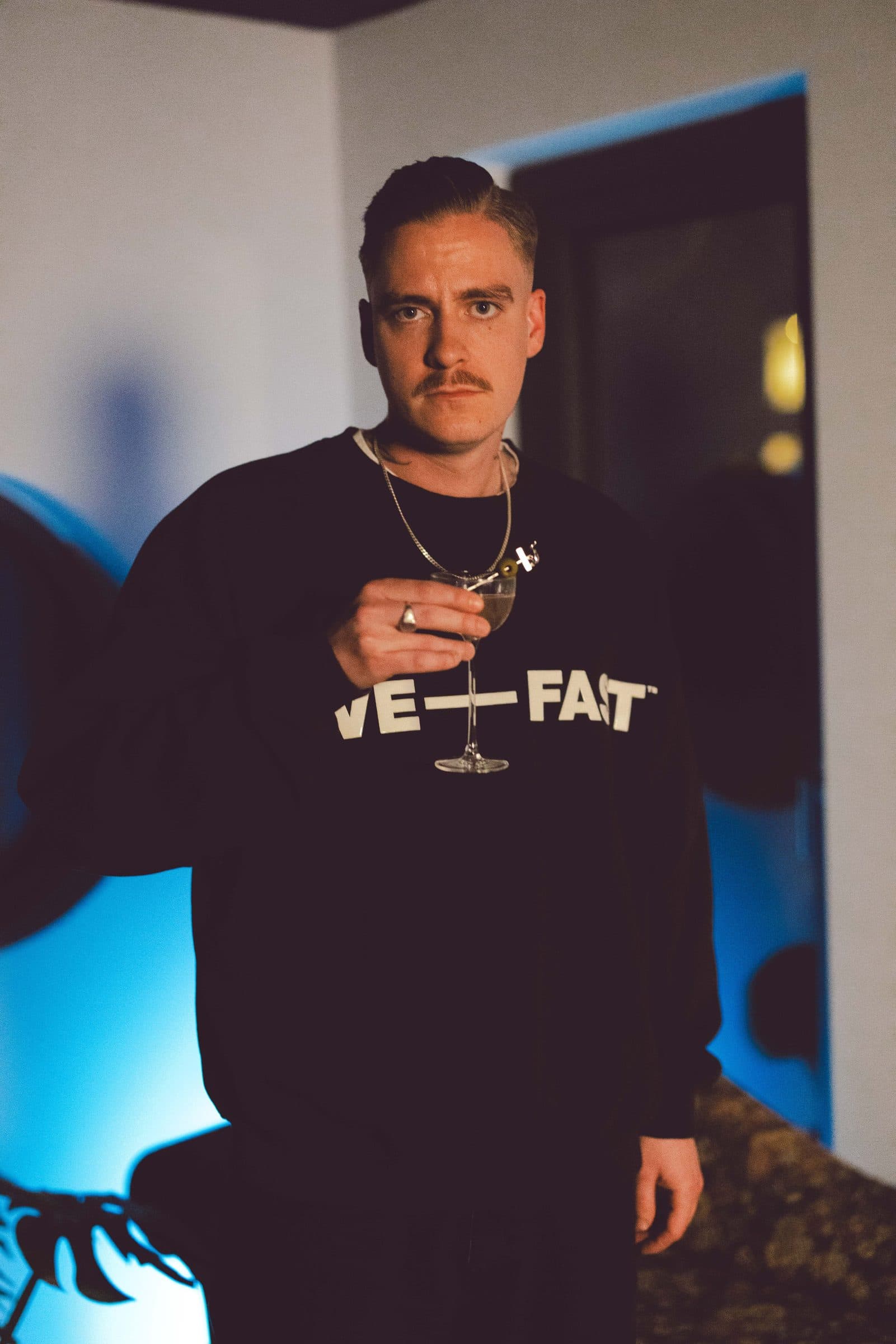 Marvin im schwarzen Sweatshirt hält eine Coupette mit hellem Cocktail und schaut in die Kamera, links blaues Licht im Studio