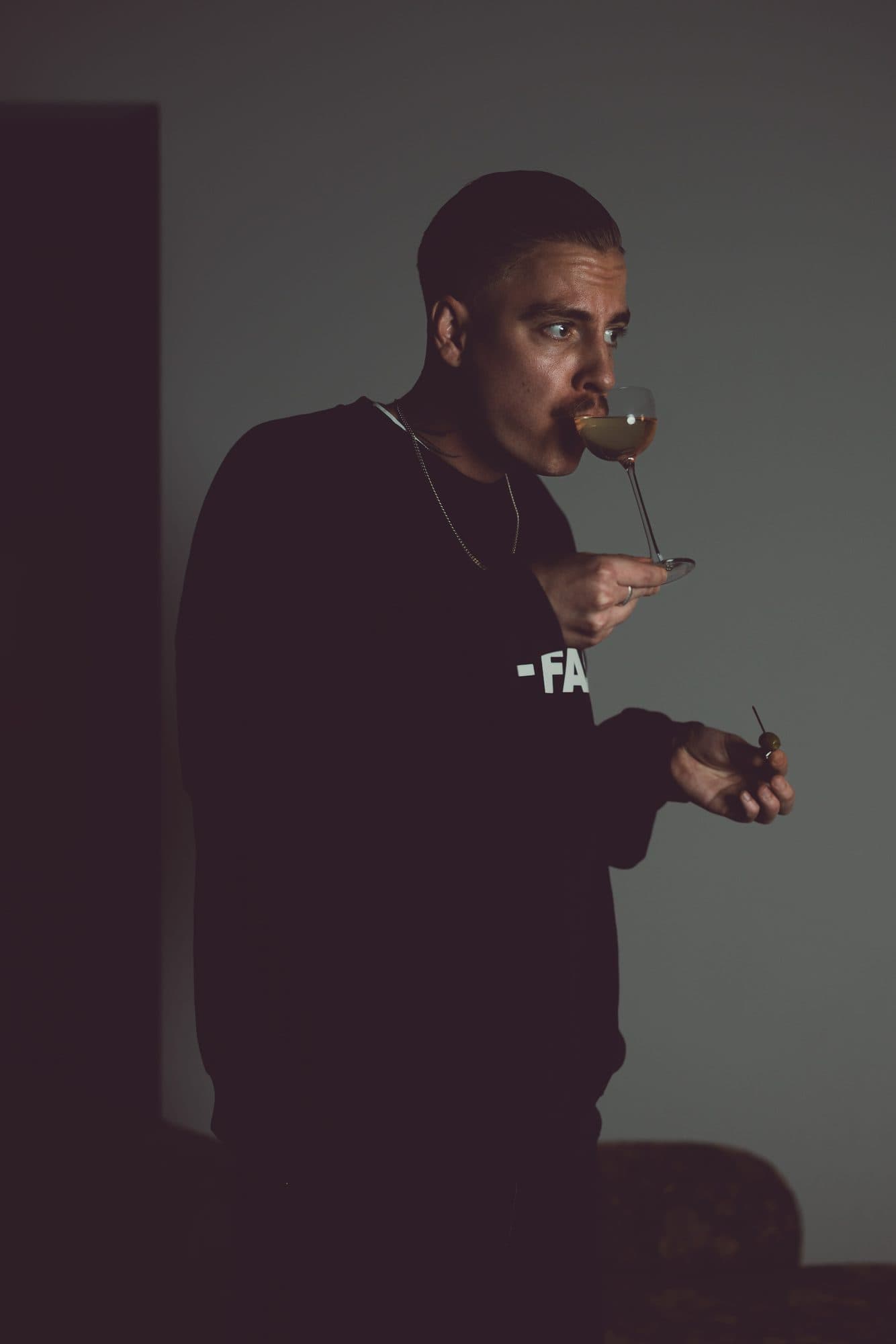 Marvin, Head Bartender von ROMI, trinkt aus einem Stielglas und hält einen Cocktailspieß mit Olive, vor grauer Wand